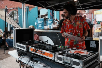 A summery-looking DJ is looking through his vinyl records while performing at Underfall Yard, a outside bar is behind him selling drinks during the festival.