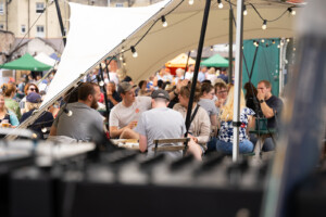 Outdoor cafe scene with a crowd seated under a canopy at Underfall Yard. People are chatting and enjoying food. String lights create a lively, festive atmosphere.