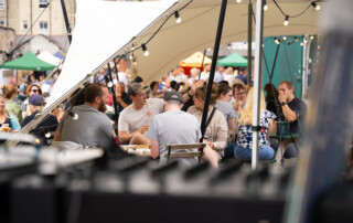Outdoor cafe scene with a crowd seated under a canopy at Underfall Yard. People are chatting and enjoying food. String lights create a lively, festive atmosphere.