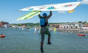 A man jumping into Bristol Harbour during the Bristol Birdman event. It is a sunny summer day with other vessels sailing in the background, with Underfall Yard spotted in the distance