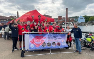 The winning team of the Dragon Boat Racing tournament celebrate their victory in front of a banner. Wearing costumes themed to The Incredibles, one team member is elevated holding the trophy and bottle of prosecco
