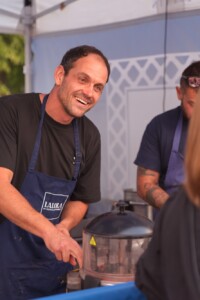 Smiling man in a blue apron cooking at an outdoor food stall. He's engaged with customers, creating a friendly and inviting atmosphere.