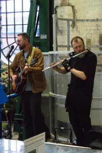 Two musicians perform indoors; one plays an acoustic guitar and sings, while the other plays a flute. The setting is casual and creative.