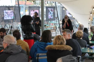 Audience seated under a tent watches one performer singing. Behind them, photos of boatbuilding workshops are displayed on the walls. The scene is lively and engaging.