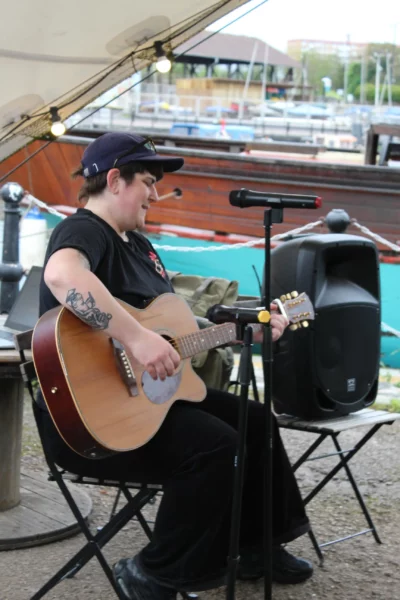 A person sits under a canopy by a waterfront, playing an acoustic guitar and singing into a microphone during an Open Mic. The setting feels casual and relaxed.