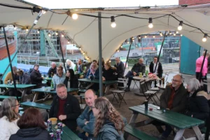 A lively group of people sitting at green picnic tables under a tent with string lights, chatting and enjoying drinks near the bustling Bristol harbour.