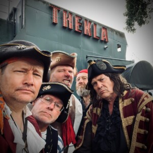 Group of men in historical pirate costumes posing, standing before Thekla, a Bristol venue and dark ship with lights.