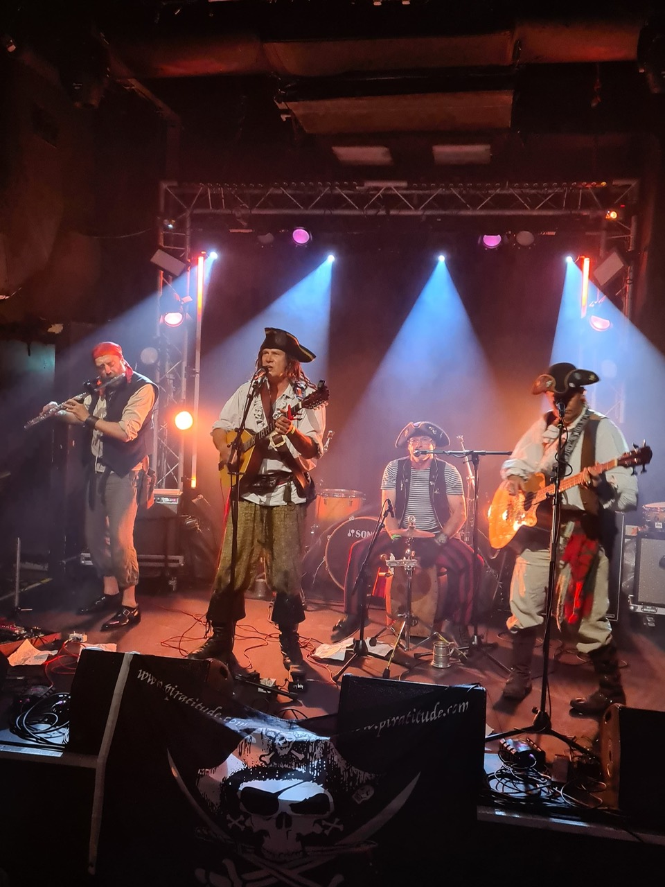 Pirate-themed rock band performing on stage with guitars, microphones, drum kit, colourful spotlights, live lights