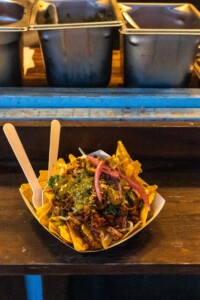 A plate of loaded nachos with shredded meat, green salsa, pickled onions, and herbs on a wooden counter. Metal trays in the background suggest a casual dining setting.