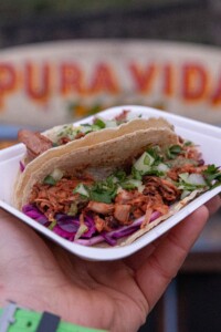 Close-up of a hand holding a tray with two tacos filled with shredded meat, cilantro, and red cabbage. A blurred "Pura Vida" sign is in the background.