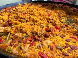 A large pan of vibrant paella filled with golden rice, pieces of chicken, red bell peppers, green beans, and slices of chorizo. The rich colours evoke warmth and flavour.