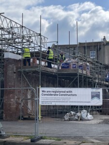 Construction site with workers in yellow vests on scaffolding, laying bricks to reinstate the Big Shed maritime workshop. A sign reads