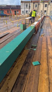 Construction workers on a wooden rooftop, discussing over a green beam and brick structure, with yellow safety markers in the background.