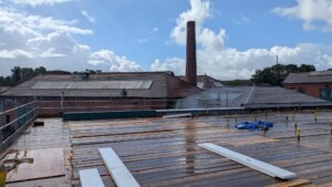 View from a rooftop showing the wall plates at Underfall Yard. Wooden planks, scaffolding, and a brick chimney are under a partly cloudy sky.