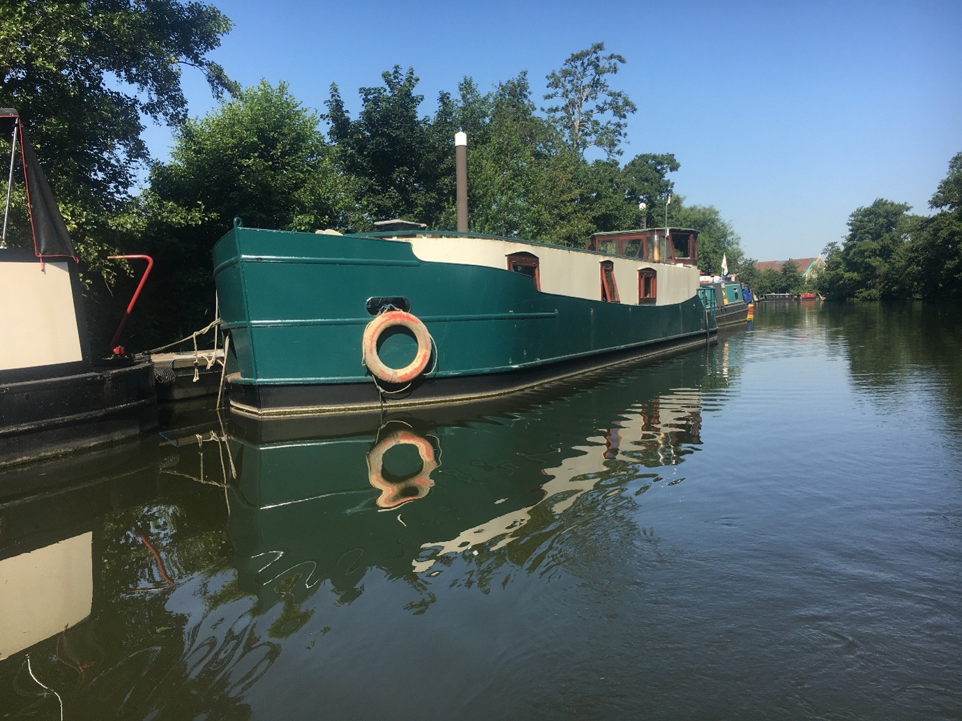 Green and white house boat moored beside dock, calm river reflecting trees and sky.