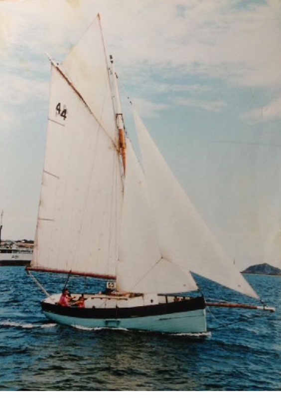 A small sailboat with the number 44 on its sail moves across calm blue water under a partly cloudy sky.