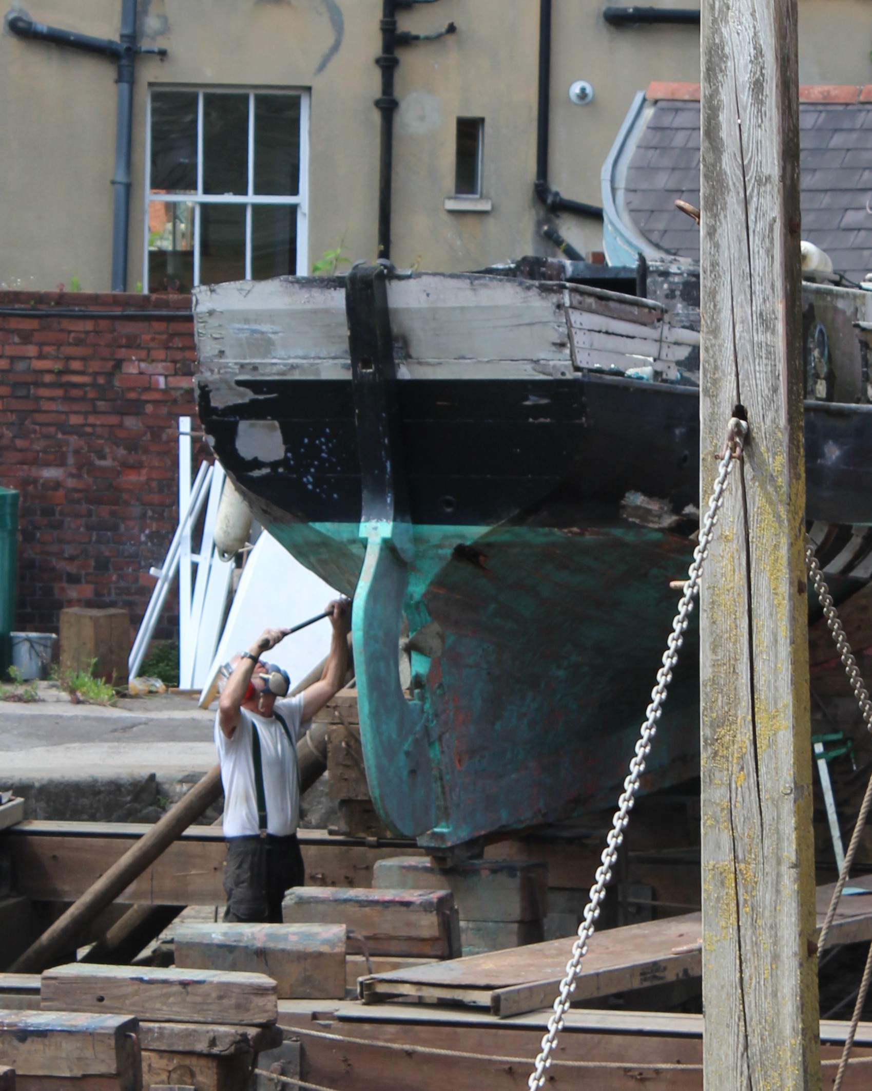 A man wearing a mask works on the hull of a burnt wooden boat in a boatyard. It is docked on the slipway in Underfall Yard