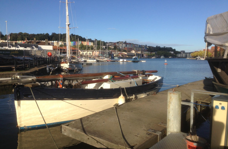 A small boat docked at Underfall Yard with colourful buildings in the background and clear blue skies.