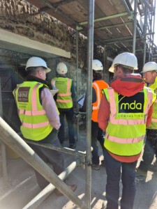 Construction workers in hard hats and high-visibility vests discussing plans near a brick wall and scaffolding.