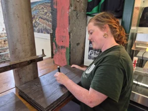 An Underfall Yard staff member smiles while writing on a slate with a marker. She wears a green shirt.The background is an industrial setting with a photo and colorful wall.