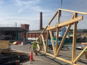 A construction worker in a high-visibility vest guides a large wooden truss by a waterfront. Scaffolding and the tall brick chimney of Underfall Yard are visible. Sunny day.
