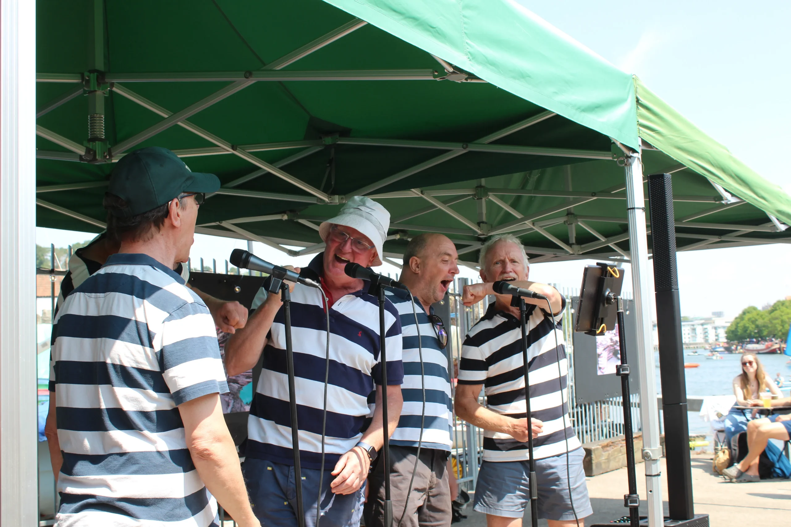Four sea shanty singers in striped shirts perform at Underfall Yard at a waterfront event, singing into microphones with lively, cheerful energy.