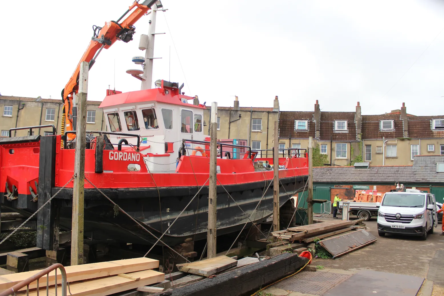 A red-and-black dredger boat rests on the slipway in Underfall Yard. Its orange crane raised beside dockside homes and a parked van; a busy, practical scene.